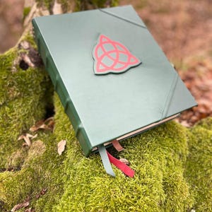 May include: A dark green leather-bound journal with a red triangular symbol on the front cover. The journal is closed and lying on a bed of green moss.