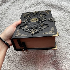 May include: Black, embossed book with gold-coloured accents. The cover features ornate celestial designs, including moons and stars. The book has a clasp closure and a black ribbon bookmark with a crescent moon charm. The corners are reinforced with gold-coloured metal.
