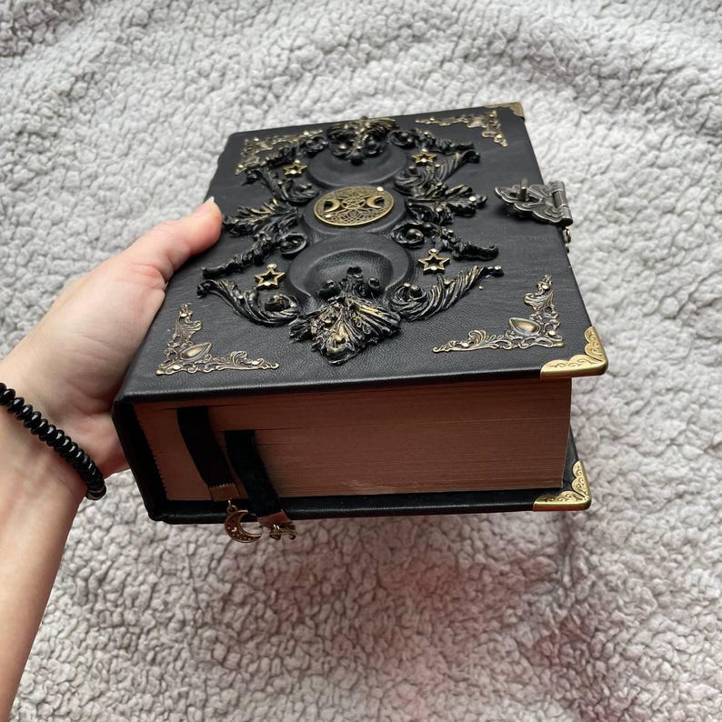 May include: Black, embossed book with gold-coloured accents. The cover features ornate celestial designs, including moons and stars. The book has a clasp closure and a black ribbon bookmark with a crescent moon charm. The corners are reinforced with gold-coloured metal.