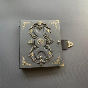 K&ouml;nnte beinhalten: Ein dunkelgraues, ledergebundenes Notizbuch mit aufwendigen goldfarbenen Metallverzierungen. Das Cover zeigt Halbmonde, Sonnen und ein zentrales Emblem. Das Notizbuch hat einen Verschluss und Eckakzente, was auf ein mystisches oder Fantasy-Thema hindeutet.
