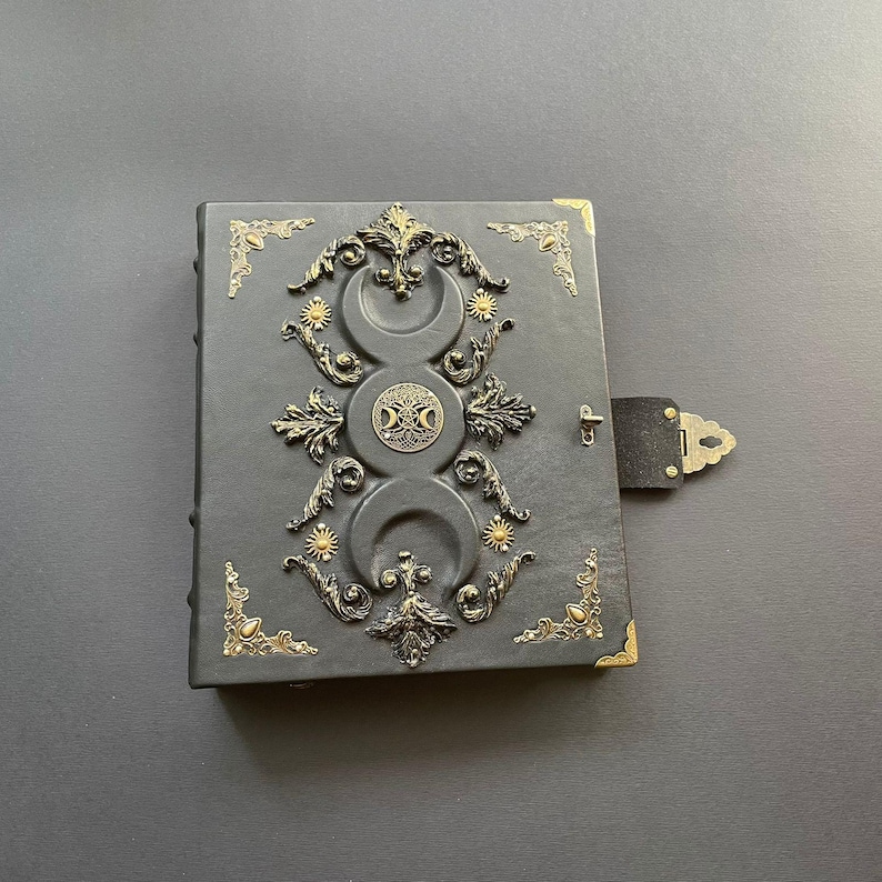 K&ouml;nnte beinhalten: Ein dunkelgraues, ledergebundenes Notizbuch mit aufwendigen goldfarbenen Metallverzierungen. Das Cover zeigt Halbmonde, Sonnen und ein zentrales Emblem. Das Notizbuch hat einen Verschluss und Eckakzente, was auf ein mystisches oder Fantasy-Thema hindeutet.