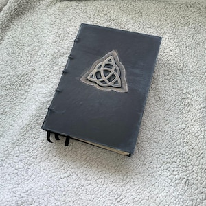May include: A black leather-bound journal with a silver-colored Celtic knot emblem. The journal has a sewn spine and a black ribbon bookmark. The cover is rectangular and has a slightly distressed appearance, suggesting a vintage or handcrafted style.