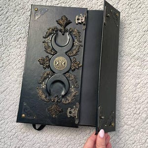 May include: A black rectangular journal with a textured cover featuring a central triple moon symbol, ornate bronze-colored filigree accents, and small star details. The journal has metal corner protectors and two side latches to secure the cover closed.