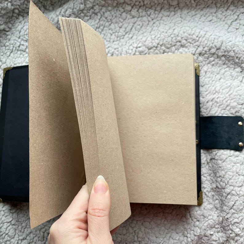 May include: A black journal with a leather strap closure and brass corner protectors. The pages are a light brown colour, and the journal is open, revealing the blank pages. The journal is on a soft, textured surface.