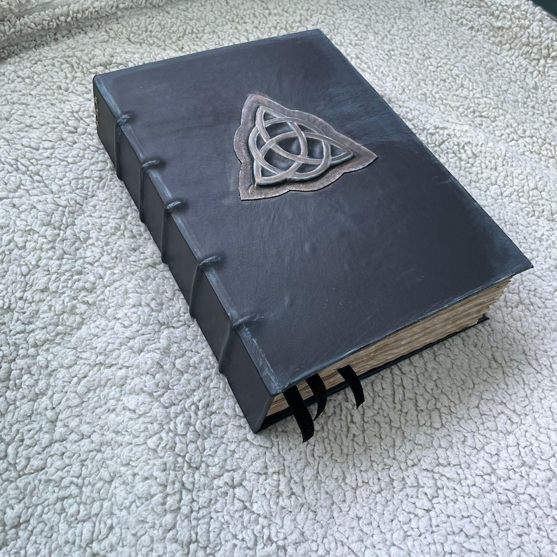 May include: A dark blue leather-bound book with a silver Celtic knot emblem. The book has a worn appearance, with visible stitching along the spine and black ribbon bookmarks. The pages have a vintage, aged look.