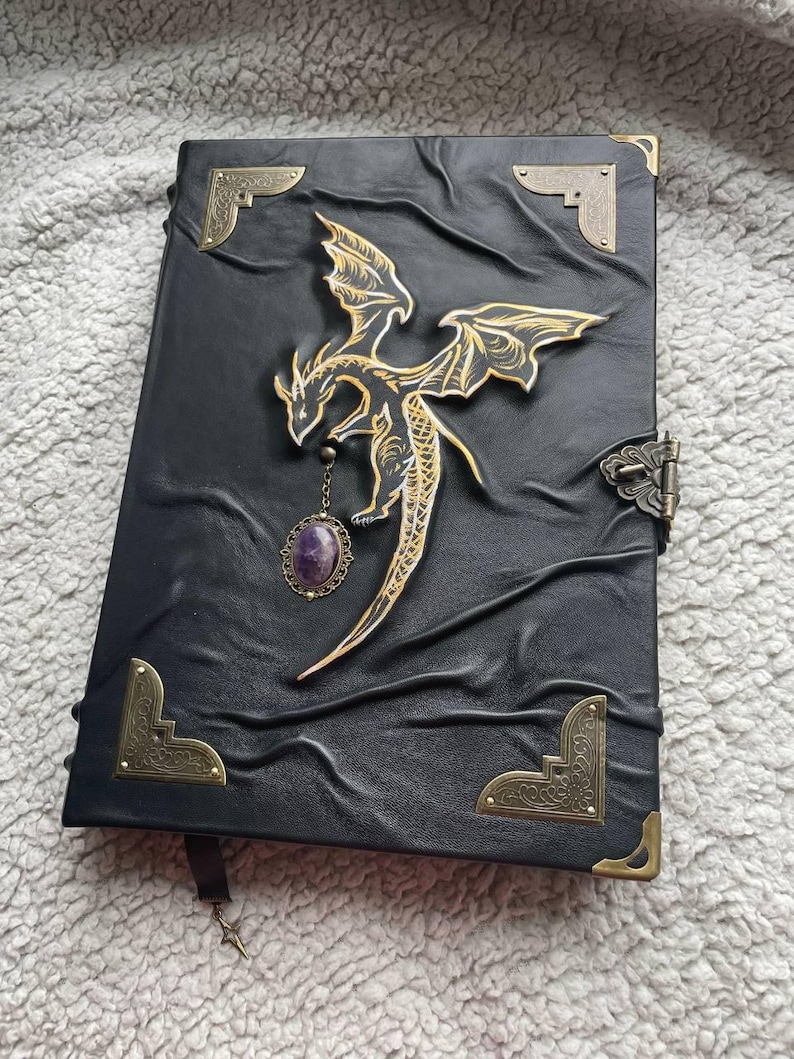 Pu&ograve; includere: Diario in pelle nera con un design a drago dorato. Il drago &egrave; impreziosito da dettagli dorati e un ciondolo con pietra preziosa viola. Il diario ha angoli decorativi e una chiusura a fermaglio. &Egrave; visibile un segnalibro con un ciondolo a stella.
