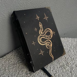 May include: Black leather journal with a gold snake and star design. The journal has a gold clasp and a black ribbon bookmark.