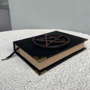 May include: Black leather-bound journal with a gold pentagram design on the cover. The journal has gold corner accents and a brown ribbon bookmark.