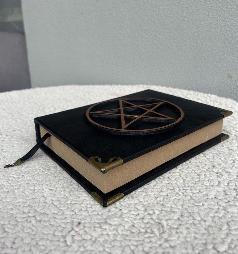 May include: Black leather-bound journal with a gold pentagram design on the cover. The journal has gold corner accents and a brown ribbon bookmark.