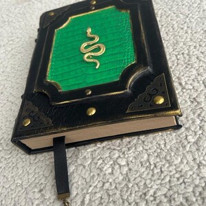 Pode incluir: Um di&aacute;rio encadernado em couro preto com uma capa verde em relevo que apresenta um design de cobra dourada. O di&aacute;rio tem um fecho met&aacute;lico decorativo e um marcador de fita preta com um chaveiro.
