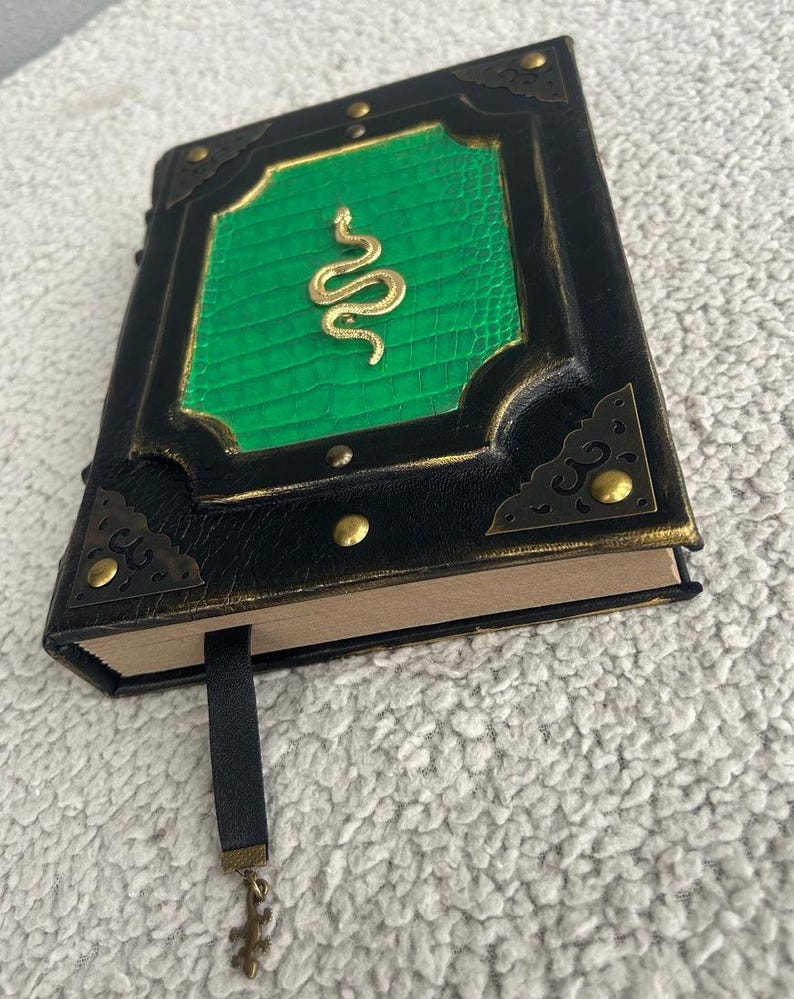 Pode incluir: Um di&aacute;rio encadernado em couro preto com uma capa verde em relevo que apresenta um design de cobra dourada. O di&aacute;rio tem um fecho met&aacute;lico decorativo e um marcador de fita preta com um chaveiro.