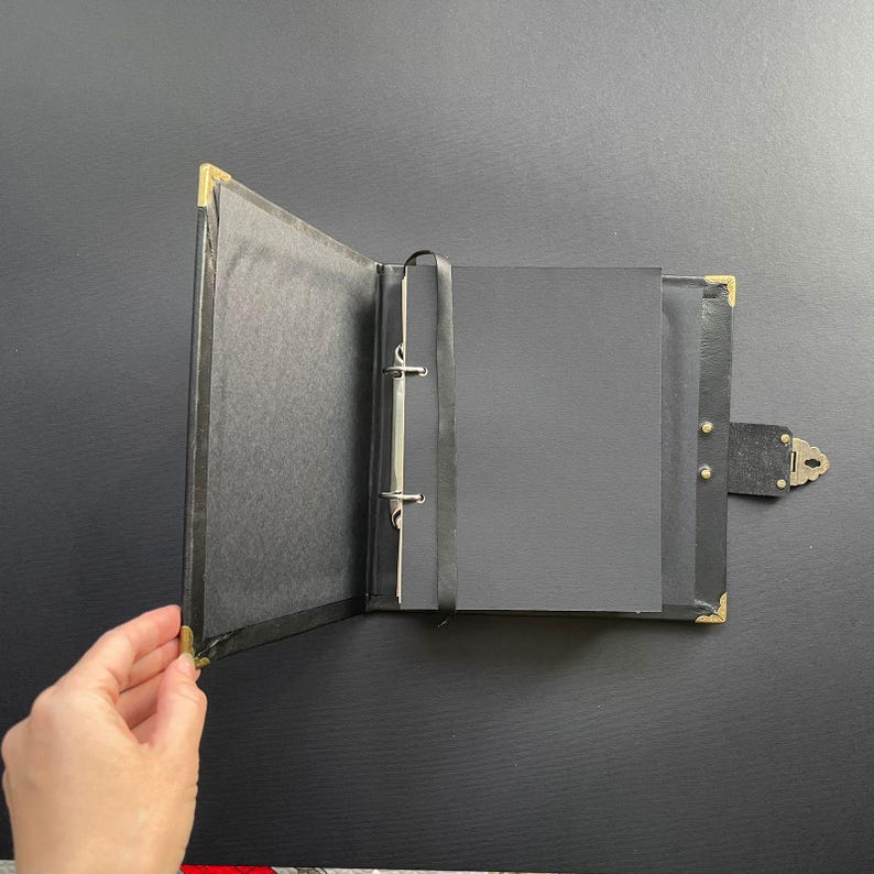 K&ouml;nnte beinhalten: Ge&ouml;ffnetes schwarzes Fotoalbum mit goldenen Eckakzenten und einem Verschluss. Das Album zeigt schwarze Seiten und einen Bindemechanismus. Es wird vor schwarzem Hintergrund gehalten. Abmessungen in Zentimetern.