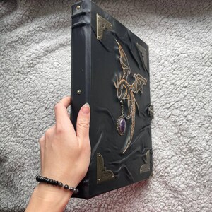 Pu&ograve; includere: Diario in pelle nera con un disegno di drago. Il diario presenta un emblema di drago dorato, angoli decorativi e un ciondolo con pietra preziosa viola. La copertina ha un aspetto strutturato e in rilievo e una chiusura in metallo.