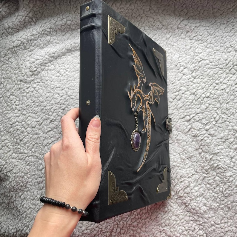 Pu&ograve; includere: Diario in pelle nera con un disegno di drago. Il diario presenta un emblema di drago dorato, angoli decorativi e un ciondolo con pietra preziosa viola. La copertina ha un aspetto strutturato e in rilievo e una chiusura in metallo.