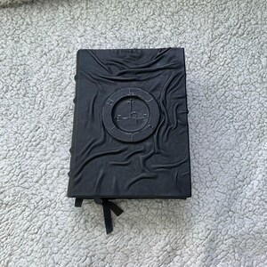 May include: A black, embossed book with a circular emblem on the cover. The emblem features a cross-like symbol within a ring. The book has a textured, wrinkled appearance and a black ribbon bookmark.