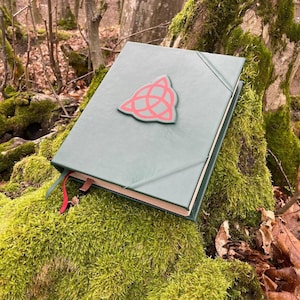 May include: A green leather-bound journal with a red triangular symbol on the front cover. The journal is lying on a bed of moss.