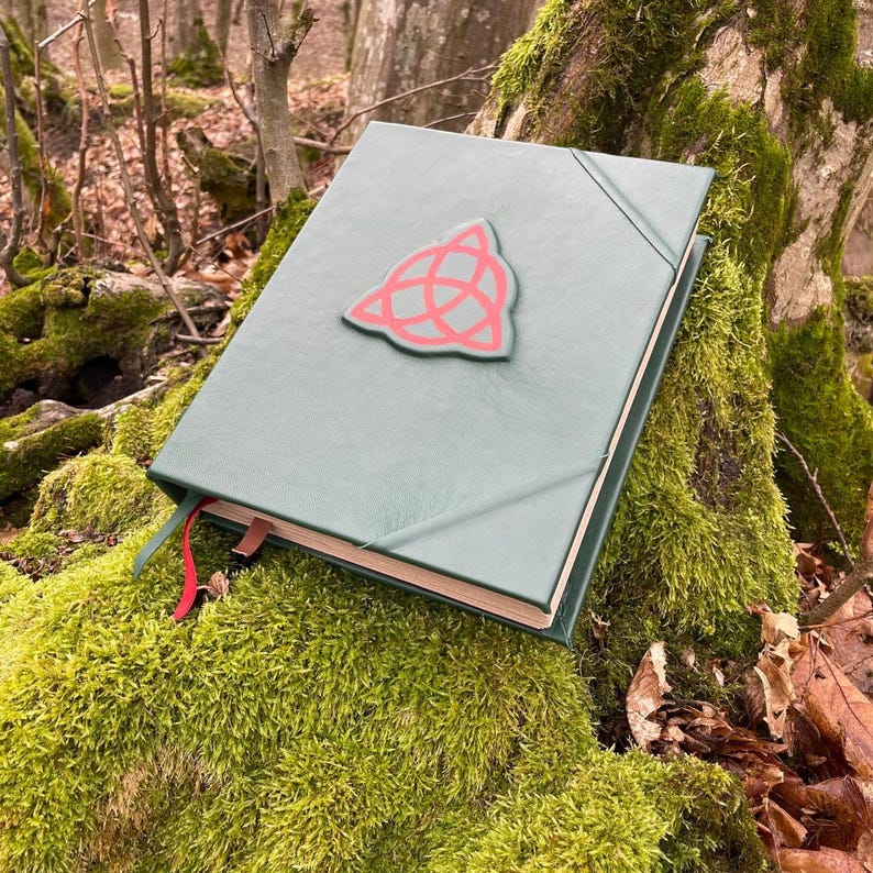 May include: A green leather-bound journal with a red triangular symbol on the front cover. The journal is lying on a bed of moss.