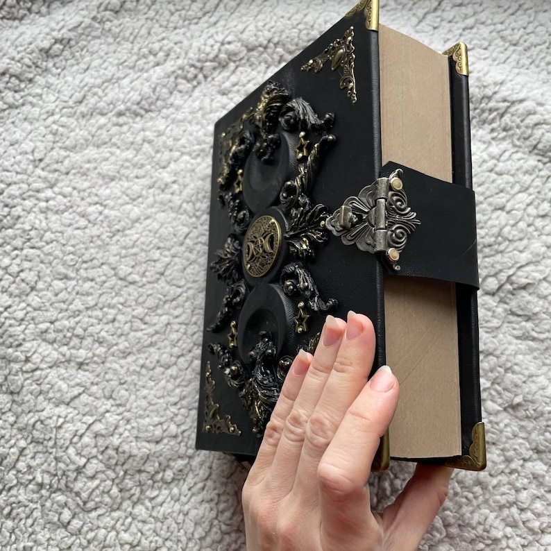 May include: A black, decorative book with gold-coloured embellishments and a clasp. The cover has detailed relief designs and corner protectors. The book is open, showing the pages.