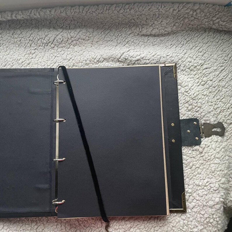 May include: Open black journal with a lock and key closure. The journal has a black ribbon bookmark and metal ring binder. The pages are black and the edges are beige. The journal is open on a textured, light gray surface.
