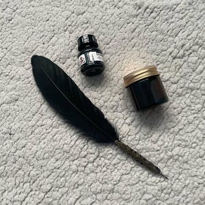 Può includere: Una penna a calamo nera con un manico in metallo decorato, una piccola boccetta di inchiostro nero con etichetta bianca e un piccolo barattolo di vetro ambrato con tappo a vite color oro. Gli oggetti sono disposti su una superficie ruvida.