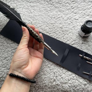 Puede incluir: Una pluma negra con un mango plateado decorativo y una pluma negra. La pluma está junto a un tintero negro y un soporte negro con puntas adicionales. También se ve una pulsera de cuentas negras.