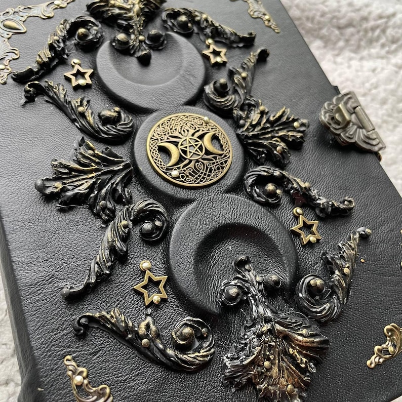 May include: Black leather journal with embossed crescent moons, stars, and ornate bronze-coloured embellishments. A central circular emblem features a triple moon symbol and a pentagram. The journal has a metal clasp.