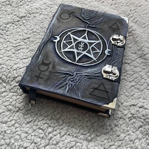 May include: A dark purple and black journal with a textured cover featuring a central silver seven-pointed star emblem, various occult symbols, two silver metal latches on the right side, and silver corner protectors on the front cover edges.