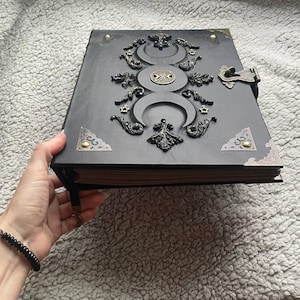 May include: A black leather-bound journal with ornate metal embellishments. The cover features a triple moon design, stars, and decorative corner accents. A metal clasp secures the journal, and a black beaded bracelet is visible.
