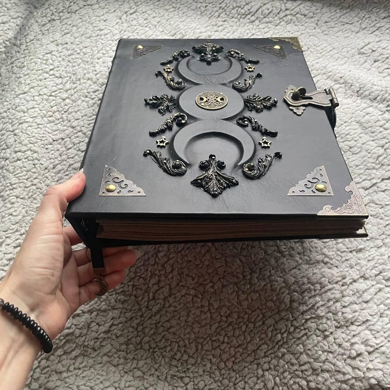 May include: A black leather-bound journal with ornate metal embellishments. The cover features a triple moon design, stars, and decorative corner accents. A metal clasp secures the journal, and a black beaded bracelet is visible.