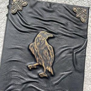 Pu&ograve; includere: Diario in pelle nera con un design a corvo dorato. Il diario ha una superficie strutturata e dettagli decorativi agli angoli. Il corvo &egrave; in rilievo sulla copertina, con piume dettagliate e una posa in piedi. Un taccuino unico e artistico.