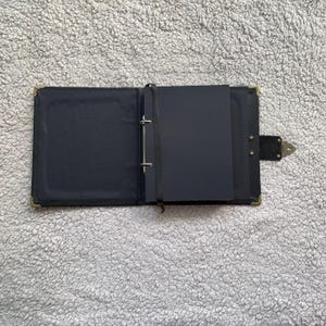 K&ouml;nnte beinhalten: Ge&ouml;ffnetes schwarzes Fotoalbum aus Leder mit goldenen Ecksch&uuml;tzern und einem Verschluss. Das Album hat schwarze Seiten und ein Leseband. Das Album ist ge&ouml;ffnet und zeigt die Innenseiten und Binderinge.