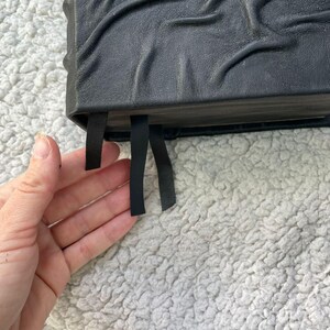 May include: A black leather-bound journal with embossed detailing. The journal has three black ribbon bookmarks. The cover is textured and the pages are visible. The journal is on a white, fluffy surface.