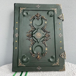 May include: A dark green leather-bound journal with intricate gold and silver embellishments. The cover features a triple moon symbol and other decorative elements.