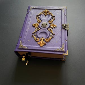 Könnte beinhalten: Ein purpurfarbenes, ledergebundenes Tagebuch mit einem dekorativen Gold- und Silberdesign, das drei Mondsicheln und einen keltischen Knoten zeigt. Das Tagebuch hat einen Metallverschluss und ein schwarzes Band als Lesezeichen.