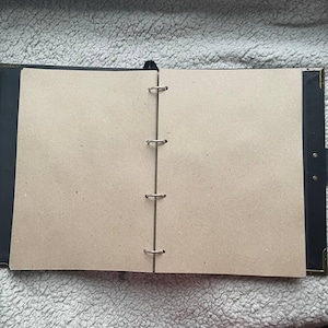 May include: Open black binder with three metal rings and blank, tan-colored pages. The binder has black corners and a black ribbon bookmark. The pages are ready to be filled with notes, sketches, or other creative content.