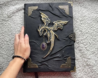 May include: Black leather journal with a gold dragon design. The dragon has outstretched wings and is embellished with a purple gemstone pendant. The journal has gold corner accents and a clasp.