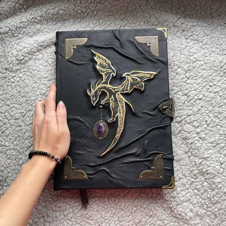 Pu&ograve; includere: Diario in pelle nera con un design a drago dorato. Il drago ha le ali spiegate e un ciondolo con pietra preziosa viola. Il diario ha angoli dorati e una chiusura.