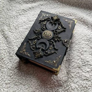 May include: Black leather journal with bronze-coloured decorative elements. The cover is adorned with crescent moons, stars, and a central circular design. Bronze accents are also on the corners and clasp, creating a gothic or celestial theme.