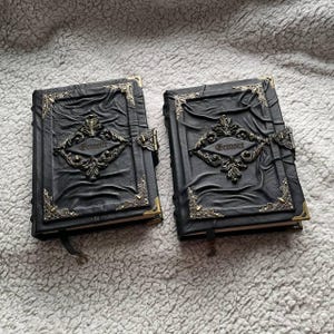 May include: Two black leather-bound journals with ornate metal clasps and corner accents. The word "Grimoire" is embossed on the front of each journal. The covers have a textured, wrinkled appearance, and each journal has a black ribbon bookmark.