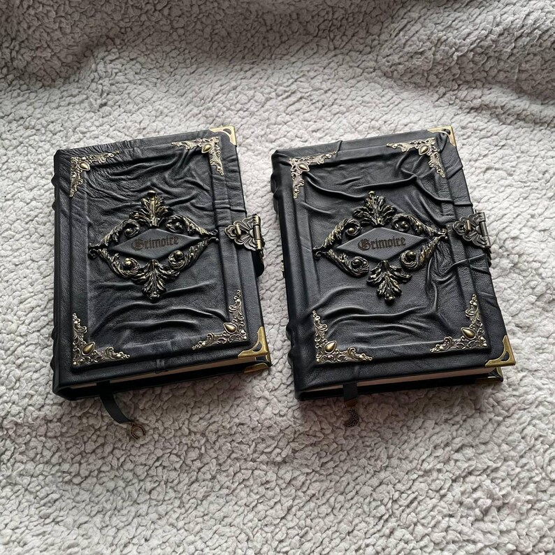 May include: Two black leather-bound journals with ornate metal clasps and corner accents. The word "Grimoire" is embossed on the front of each journal. The covers have a textured, wrinkled appearance, and each journal has a black ribbon bookmark.