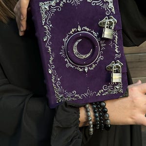 May include: A purple journal with silver detailing and a crescent moon motif. The journal features a lock closure and decorative corners. The cover is a deep purple, with silver accents providing contrast. A unique and stylish item, the journal is a great gift.