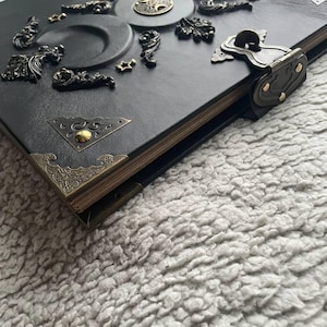 May include: A black leather-bound journal with ornate metal embellishments. The cover features a decorative lock, corner accents, and various bronze-colored metal designs, including stars and floral motifs. The edges of the pages have a golden hue.