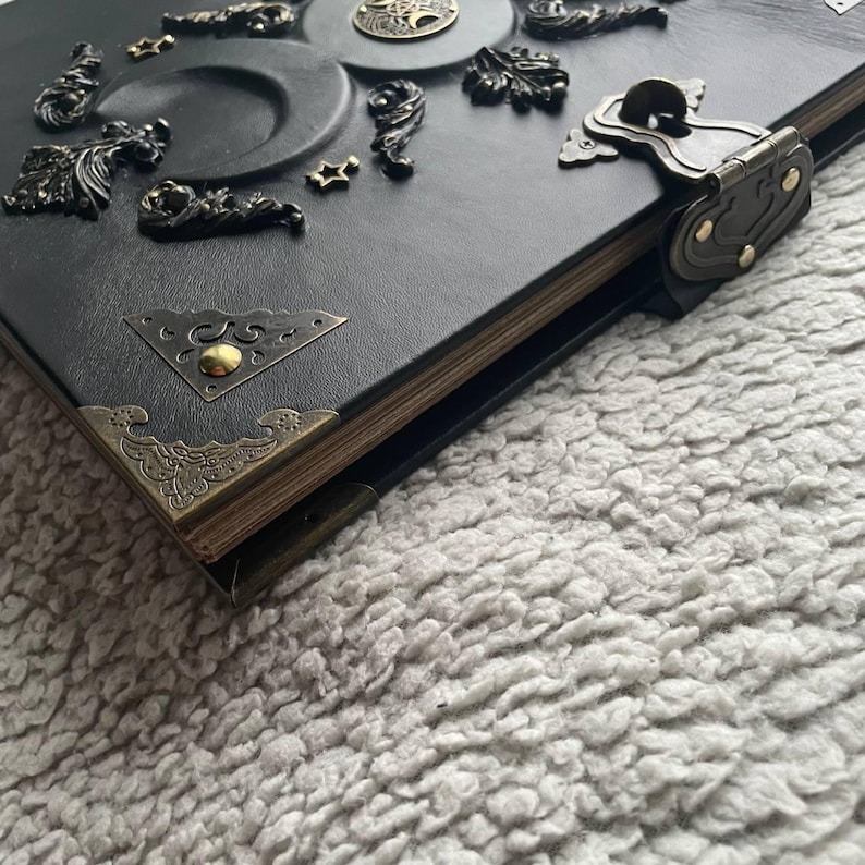 May include: A black leather-bound journal with ornate metal embellishments. The cover features a decorative lock, corner accents, and various bronze-colored metal designs, including stars and floral motifs. The edges of the pages have a golden hue.