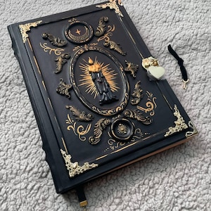 May include: A black rectangular journal featuring gold-toned metal corner protectors and a central oval relief of a lit candle. The cover is decorated with gold filigree accents, a crescent moon emblem, and a heart-shaped padlock with a small key.