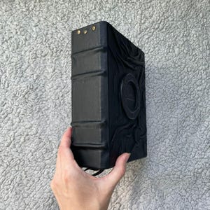 May include: A black, leather-bound book with embossed details and gold-colored rivets. The book has a circular emblem on the cover and a textured spine. The book is held by a hand.