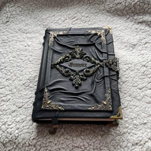 May include: Black leather-bound journal with ornate metal corner accents and a central decorative emblem. The word "Grimoire" is embossed on the cover. The journal has a clasp closure and a ribbon bookmark.