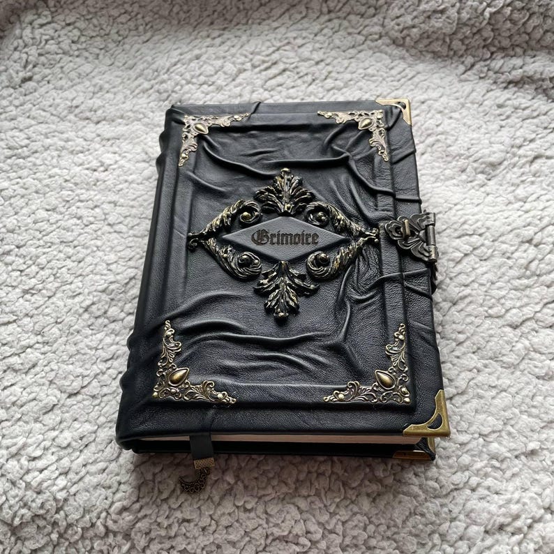 May include: Black leather-bound journal with ornate metal corner accents and a central decorative emblem. The word "Grimoire" is embossed on the cover. The journal has a clasp closure and a ribbon bookmark.