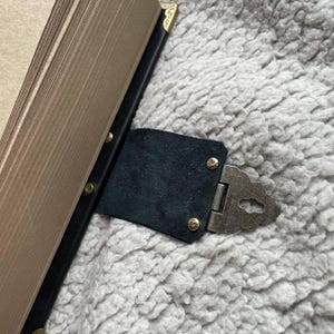 May include: A black leather book clasp with a decorative metal hinge and gold-coloured rivets. The clasp is attached to a book with gold-edged pages and black binding. The corners of the book are also gold-coloured.