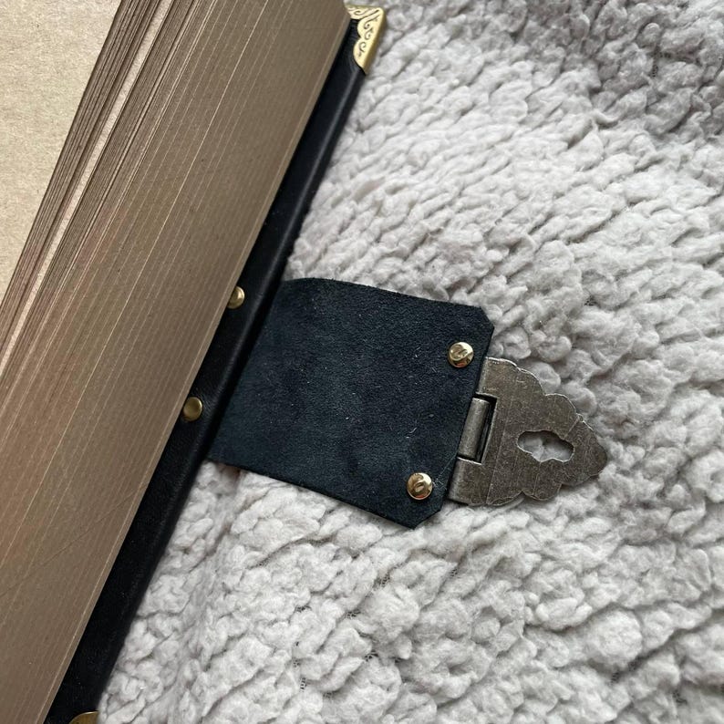 May include: A black leather book clasp with a decorative metal hinge and gold-coloured rivets. The clasp is attached to a book with gold-edged pages and black binding. The corners of the book are also gold-coloured.