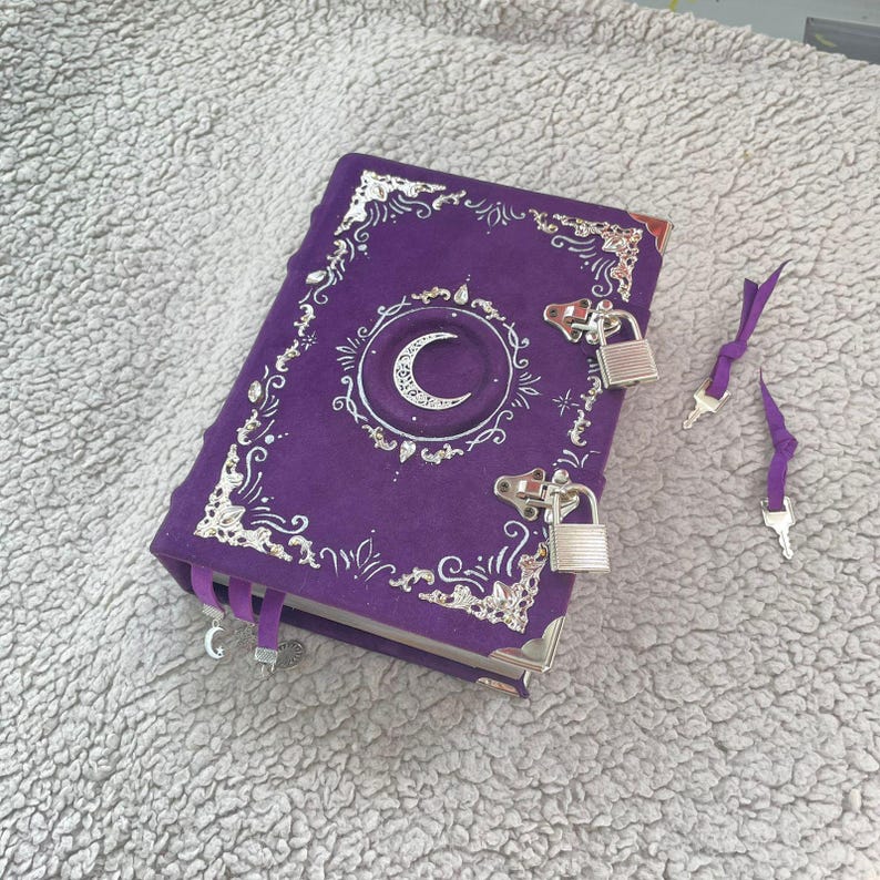 May include: A purple journal featuring a crescent moon design and silver embellishments. The journal is secured with a lock and key, with two keys and a ribbon bookmark. The cover is decorated with ornate silver filigree.